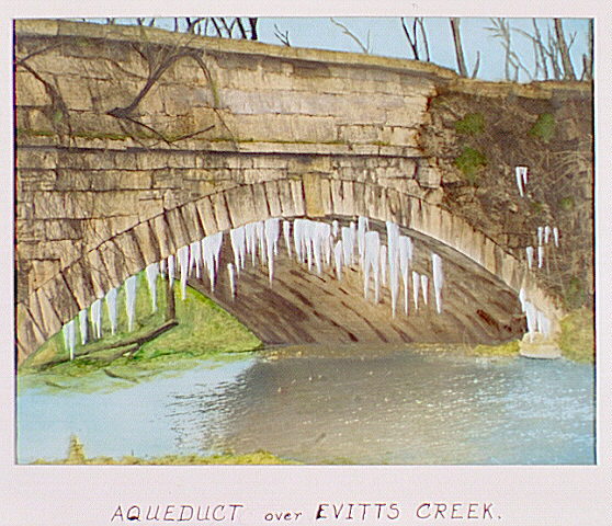 Hand-colored black-and-white photograph shows the Chesapeake and Ohio Canal’s Evitts Creek Aqueduct near Cumberland, Maryland. Note the icicles hanging underneath the aqueduct.