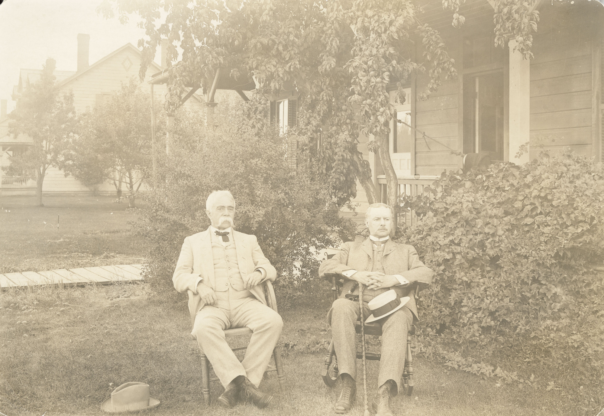Black and white photograph of two men in suits sitting in chairs in front of a house