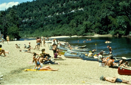 color photo with trees in background, water at right and people, boats and beach in center and left.