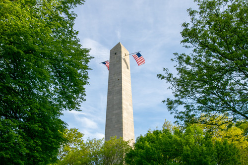 The top of a granite obelisk in morning sun, framed by green trees. Two Betsy Ross US flags hang from opposite windows beneath the pyramidion.