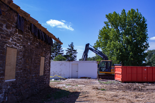 View from the Warfield house of the white garage being demolished. The brick wall of the house is in the left third of the photo, the remaining wall of the garage is in the center background with the bulldozer on its right. A large red dumpster is in the right midground and all of the buildings sit on uneven dirt.