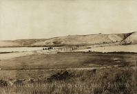 A primitive bridge structure passes over the Herring River. A horse and wagon can be seen at the top of a hill. 