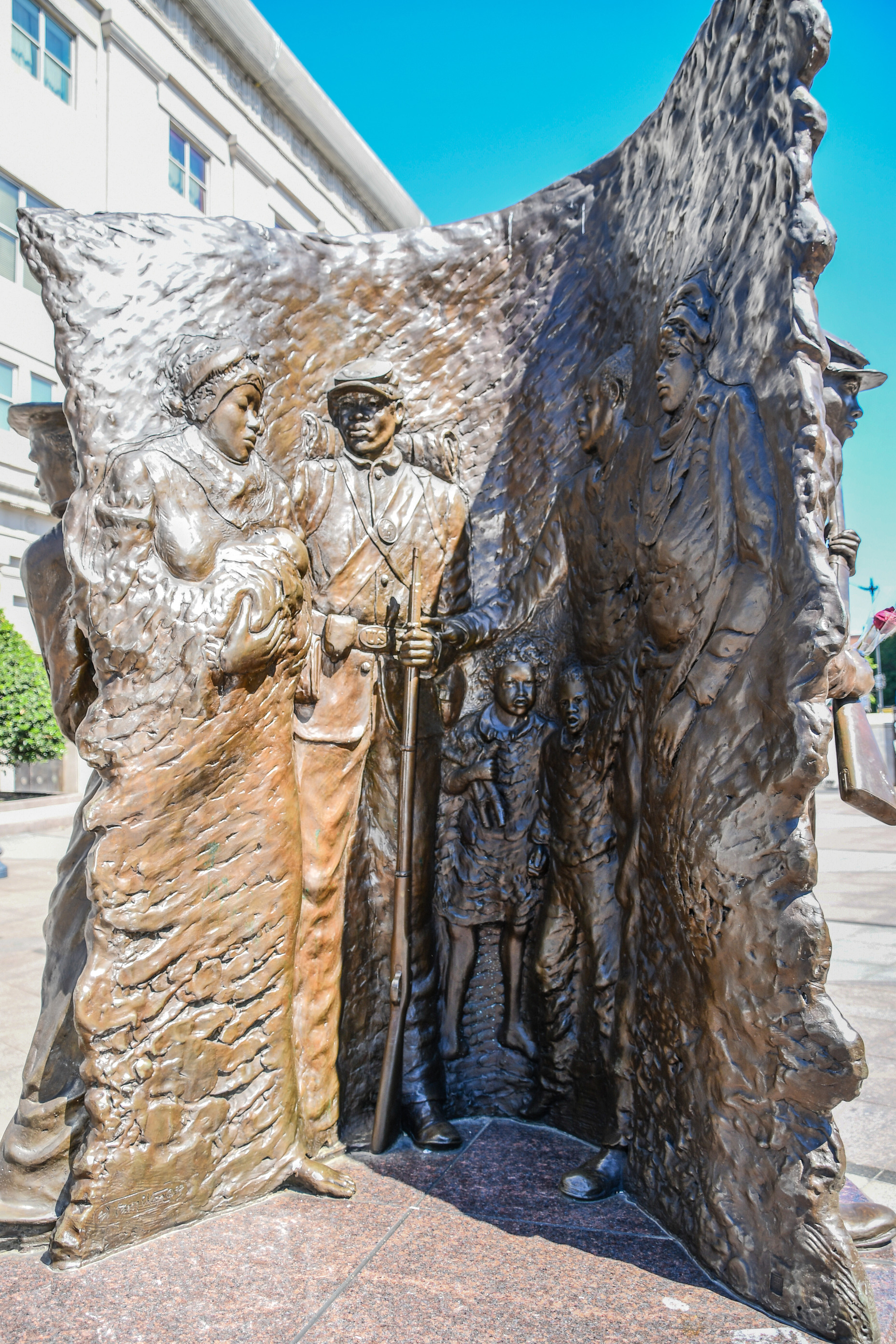 A bronze statue shows a scene of a soldier with his family; on the left the mother holding a baby in her arms, soldier looking at the baby, two small children are on the right of the soldier, and two other adults are standing to the right of the children.