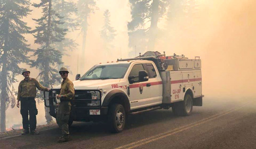 Two firefighters in yellow shirts and green pants stand in front of a white fire truck in thick wildfire smoke.