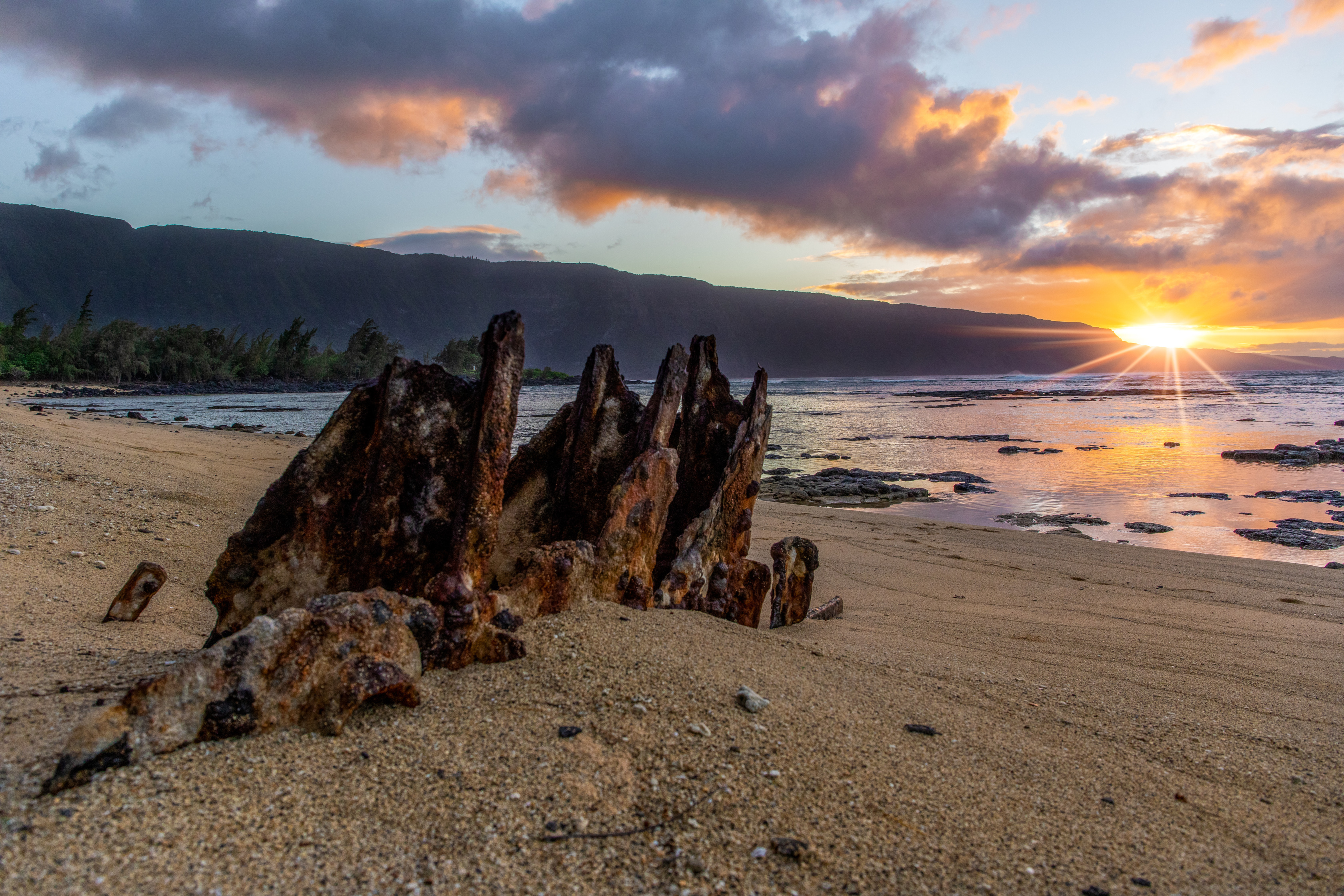 A piece of rusty metal half buried in the sand near the ocean with a setting sun in the background. 