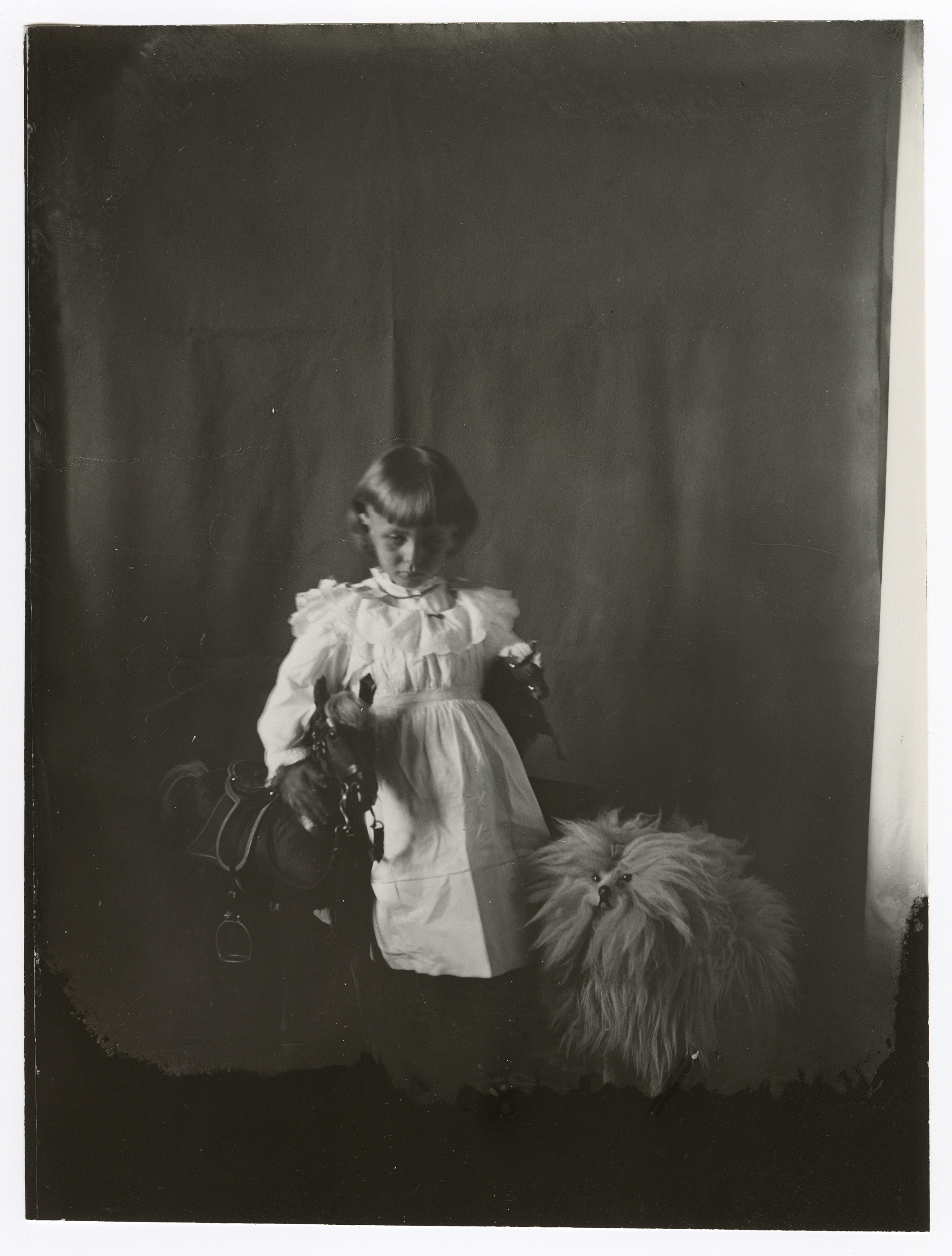 A young white girl in a white dress looks down and puts her right arm around a rocking horse and holds a horse statue in her left. Small, shaggy toy dog next to her.
