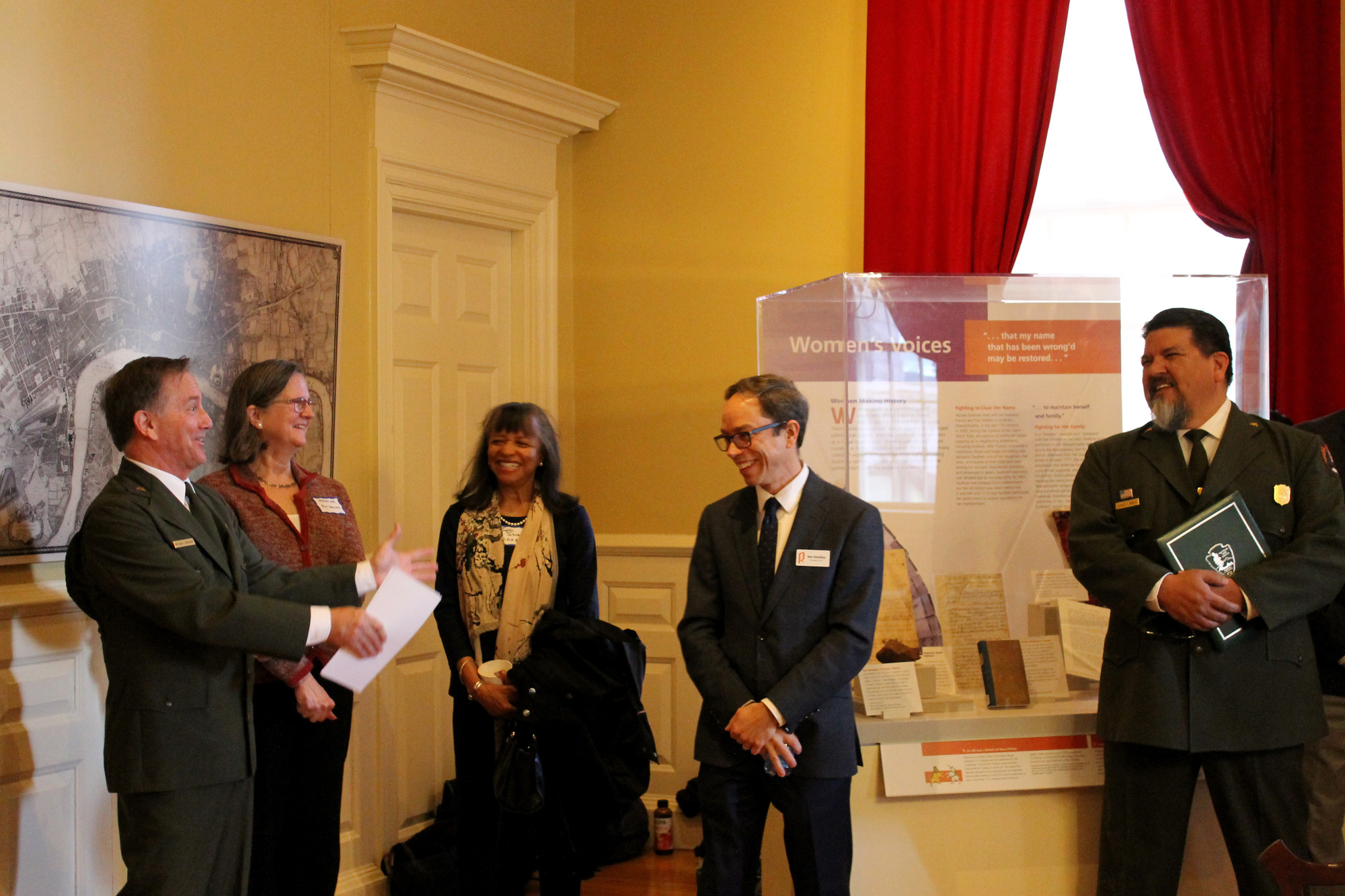 A man in a National Park Service uniform talks to a group of four people. In the back is an exhibit titled "Women's Voices. The man on the right is also in a NPS uniform and the other three people wear business clothes. 