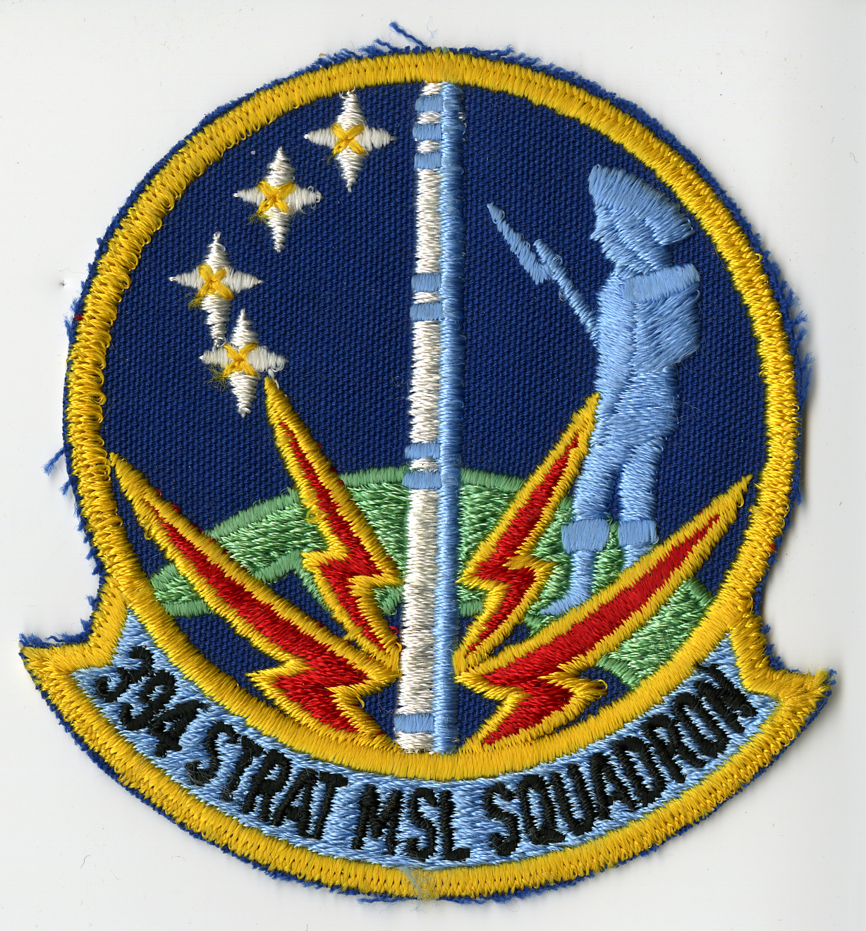 Dark blue background, circular shape, yellow border. Main patch is divided vertically by a two-tone grey missile with white four (4) point stars and yellow center on top left, bottom center is a green and blue partial sphere representing the earth with two (2) red with yellow trim lightning bolts on either side of the missile superimposed over the earth. Along right side is a grey figure resembling an 18th century Minute Man.  "394 STRAT MSL SQUADRON" in black on Bluebird Blue scroll.  Partially embroidered.
