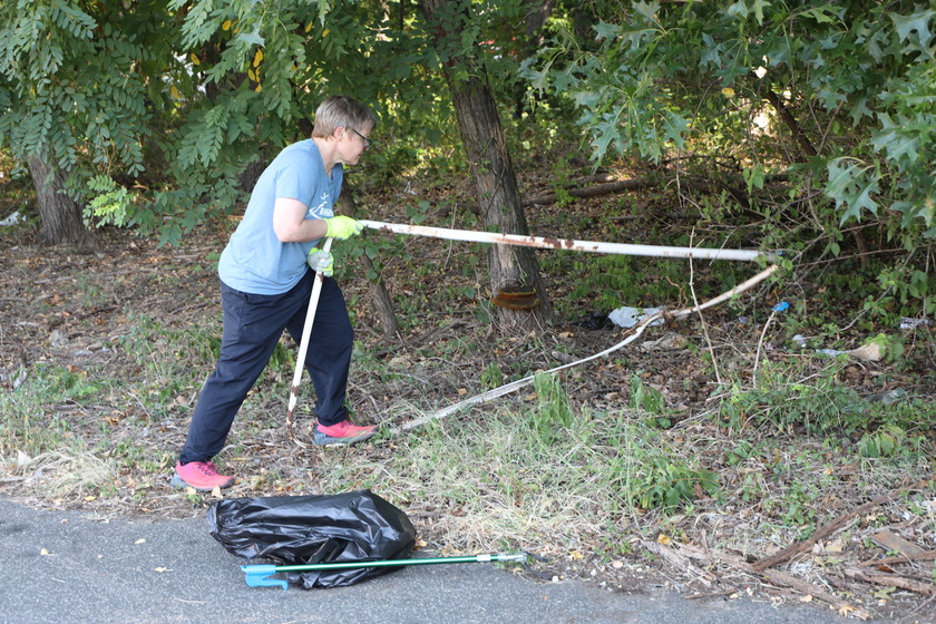 A person wearing a blue shirt, black pants, and yellow gloves pulls a bent metal frame out from beneath tree branches, with a black trash bag on the ground nearby.