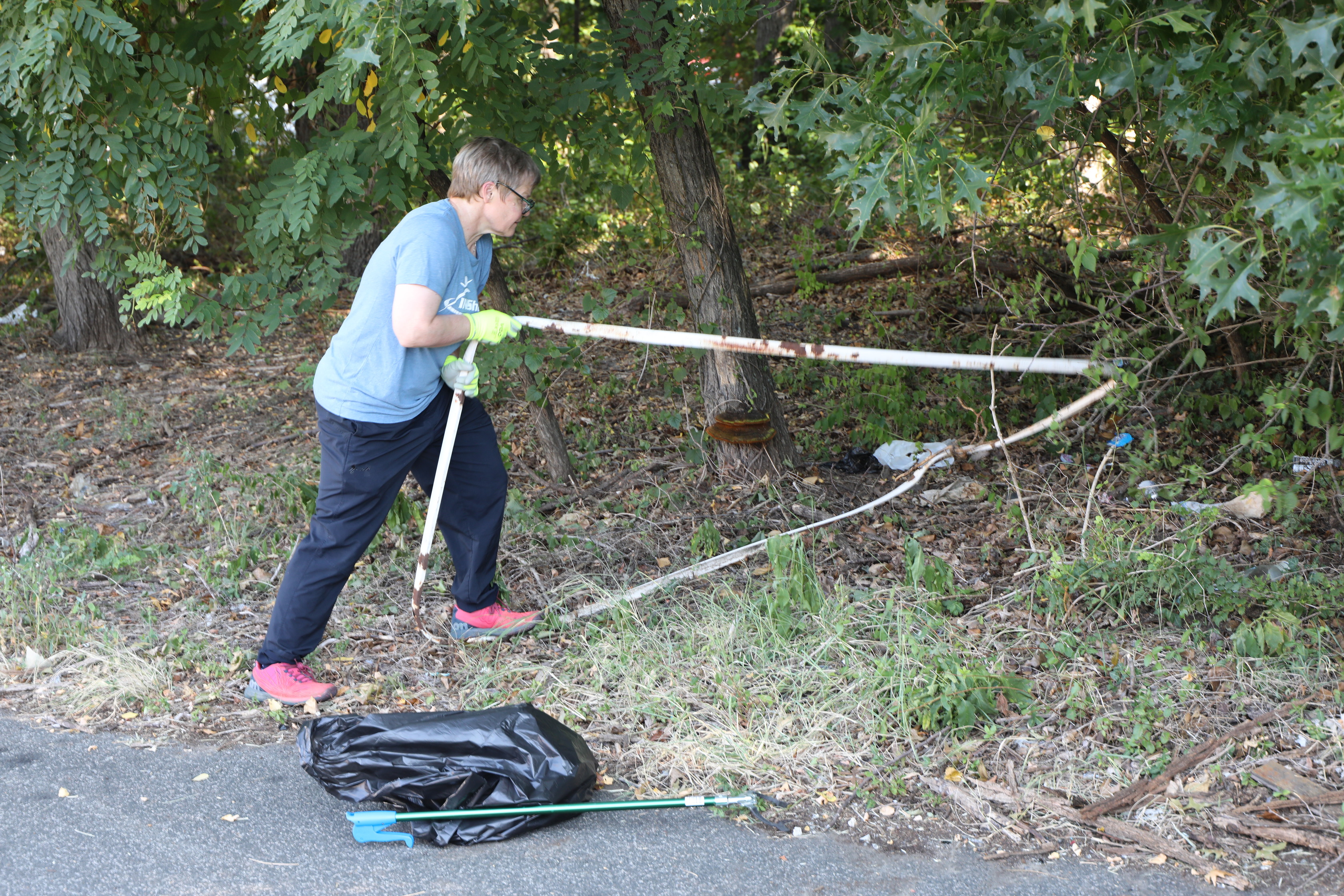 A person wearing a blue shirt, black pants, and yellow gloves pulls a bent metal frame out from beneath tree branches, with a black trash bag on the ground nearby.