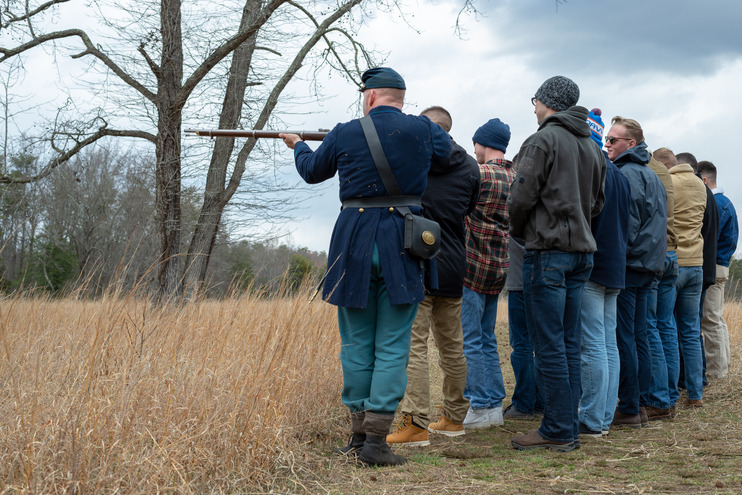 Two lines of men, seven men deep, stand facing away while a man in a Civil War Union uniform instructs them in firing weapons.