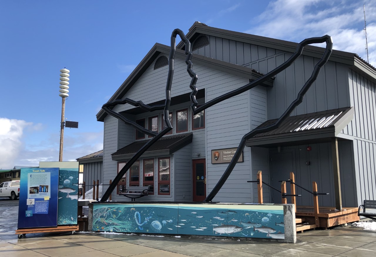 The Kenai Fjords visitor center with marine debris display out front. The display features a life-sized metal scultpture of a humpback whale with colorful mural painted on a low wall beneath the sculpture. To the side is a display on wheels with interpretive panels.