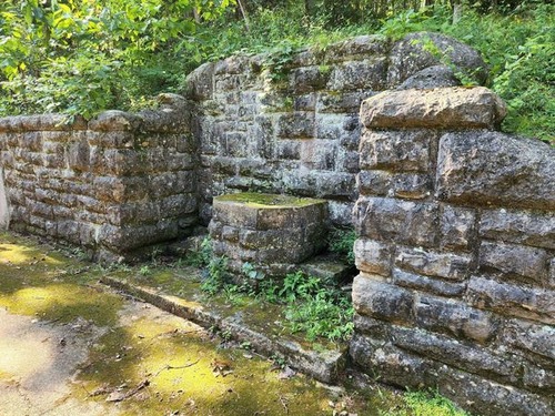 A grey stone retaining wall covered in patches of dark green moss, lighter green lichens, and dark forest algae is set into a hillside and runs from middle left to bottom right, with an aged, moss-covered concrete pad filling the space in front of it. Centered in the frame, a section of the wall is inset deeper into the hillside and has a higher top that slopes down to the level of the rest of the wall on the sides. Within the inset is a similarly constructed stone drinking fountain with a concrete basin built into the back of the inset. It has steps to either side for children and a concrete pad slightly higher than the one in front of the wall fills the inset and extends several inches in front of the wall. Low-growing forest floor plants have taken root in the moss and soil around the fountain.