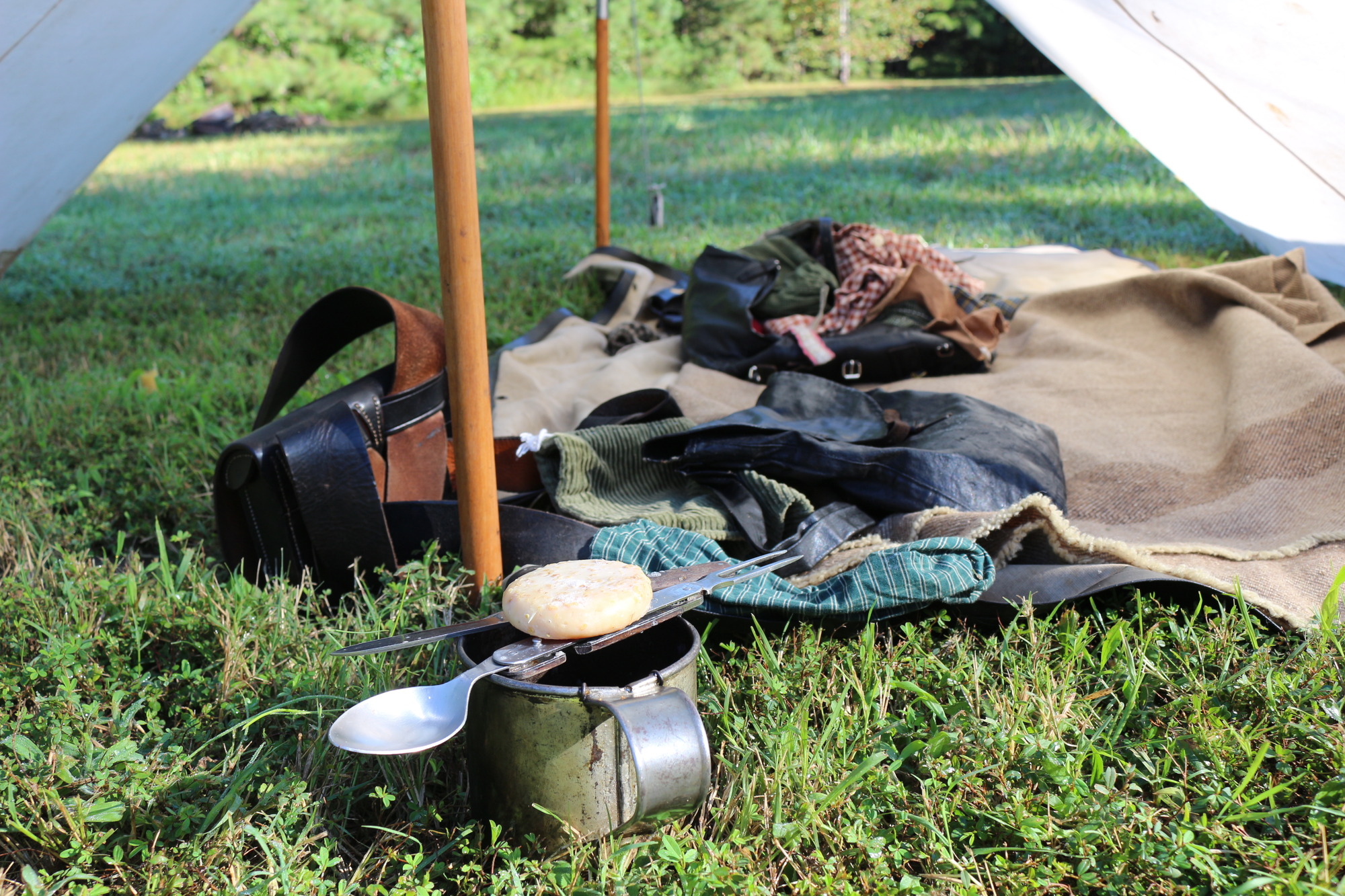Clothing and accouterments are on a blanket inside a tent. In the foreground a piece of hardtack sits on top of a fork -spoon combination tool across the brim of a tin cup.