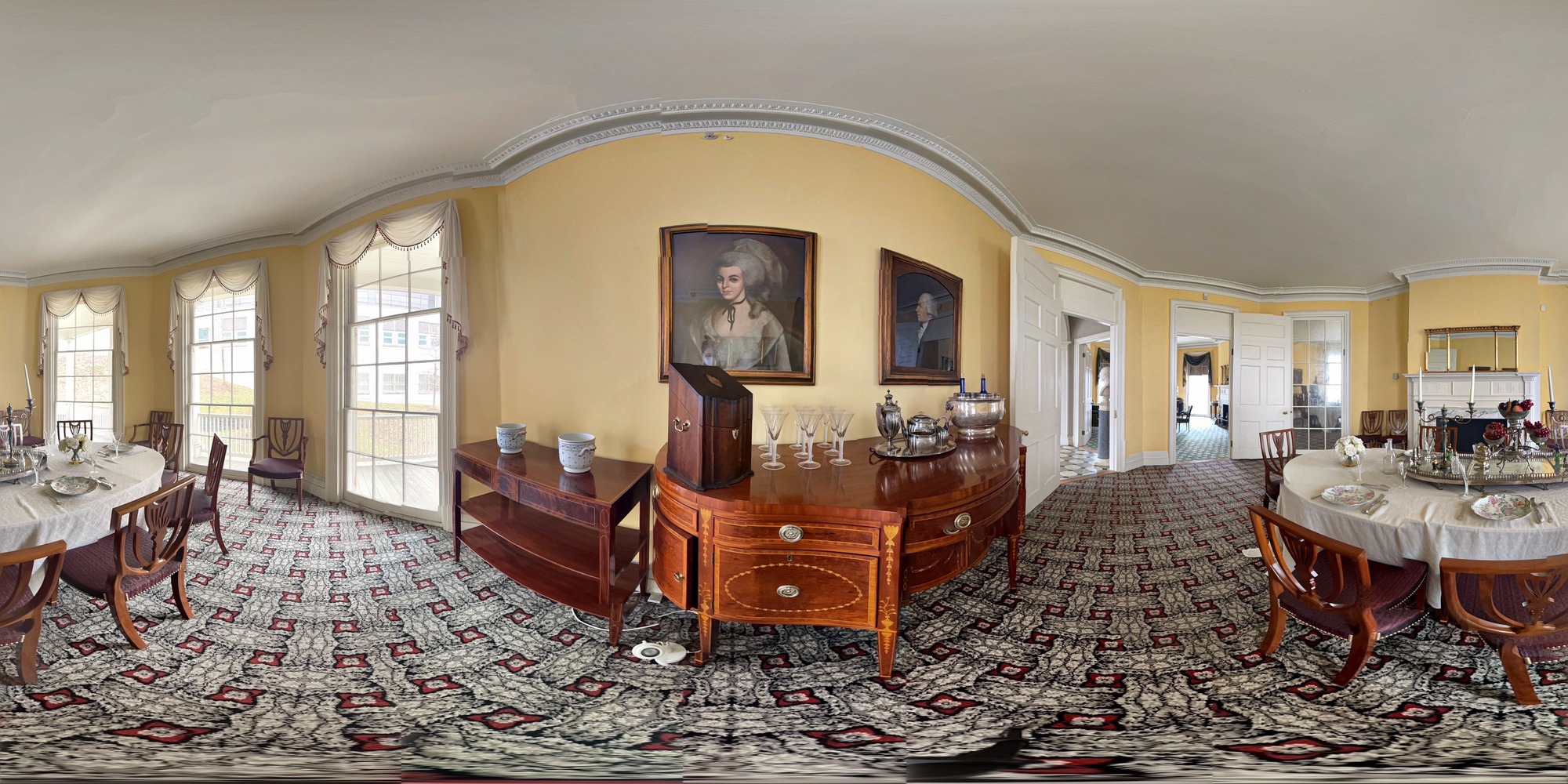 The Hamilton Grange Dining Room. Featured in this room is a large table draped in a white tablecloth. On the table are plates, silver wear, glasses, and a silver mirrored island. Atop that are candle sticks, flowers, and a centerpiece adorned with grapes. On one side of the room are three triple hung floor-to-ceiling windows, draped with dainty white curtains. Across the room are three large doors. On one wall, there are two painted portraits: one of Elizabeth, and the other, of Alexander Hamilton. Below these portraits are two serving tables, holding porcelain pots, a silver wear case, glasses, tea ware, and a wine cooler. Across from this wall is a mantle atop which is a mirror. 
