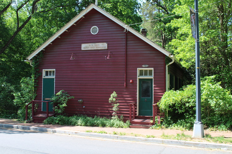 A dark red clapboard-sided building with two doors on the front. Each door has a couple of steps leading up to it. The doors are green, each with a three-pane transom window above it. A sign with the name of the building is high on the exterior wall, above and about halfway between the doors. A large bush is grown to the right of the building, obscuring the view of the windows along the side of the building.