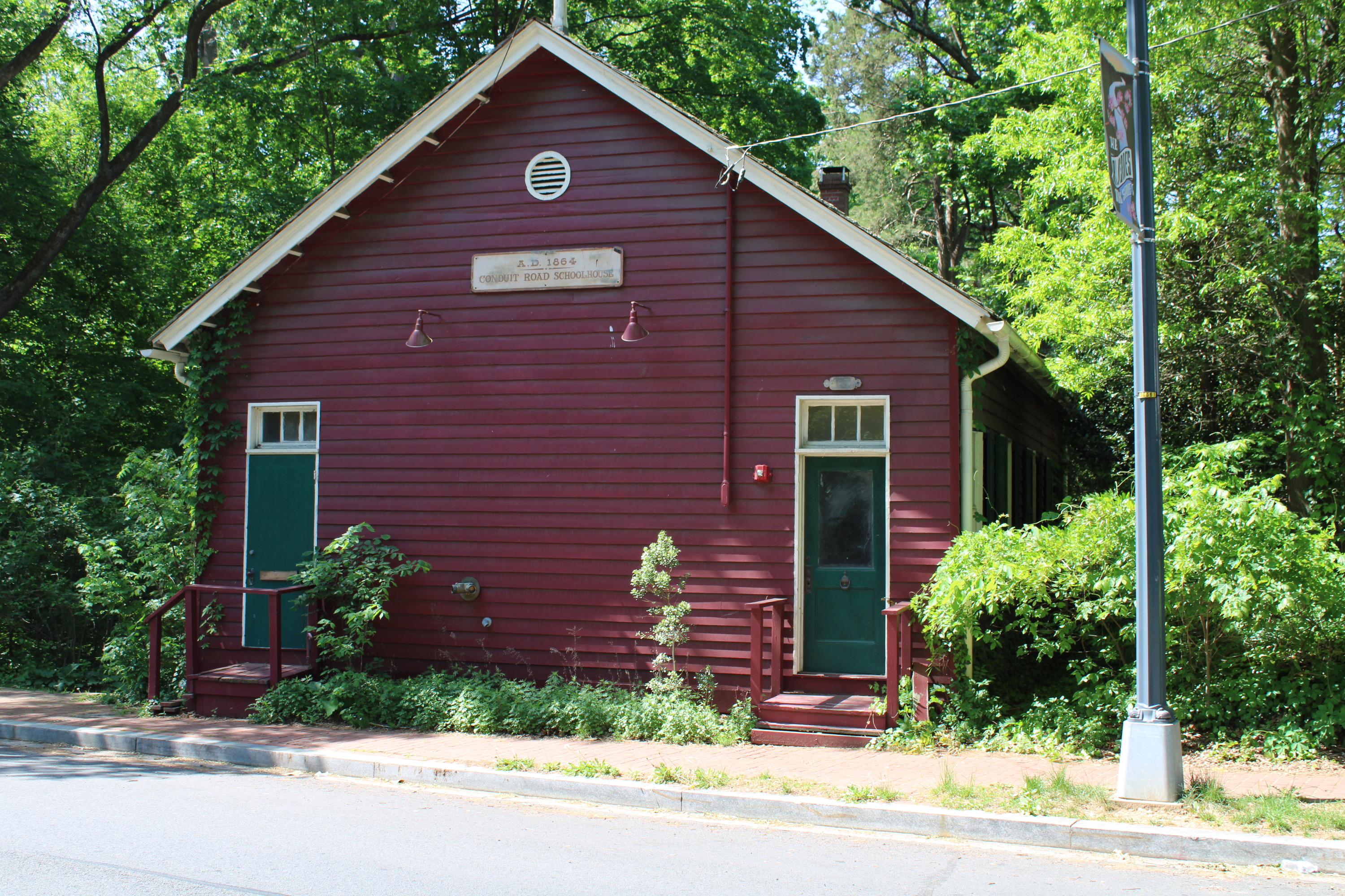A dark red clapboard-sided building with two doors on the front. Each door has a couple of steps leading up to it. The doors are green, each with a three-pane transom window above it. A sign with the name of the building is high on the exterior wall, above and about halfway between the doors. A large bush is grown to the right of the building, obscuring the view of the windows along the side of the building.