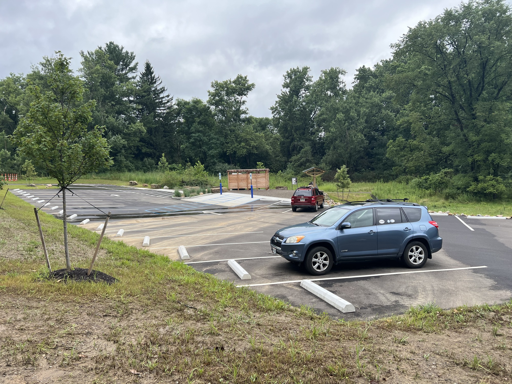 Two cars are parked in a paved lot with newly planted trees, angled spaces, a kiosk, and a wooden restroom structure.