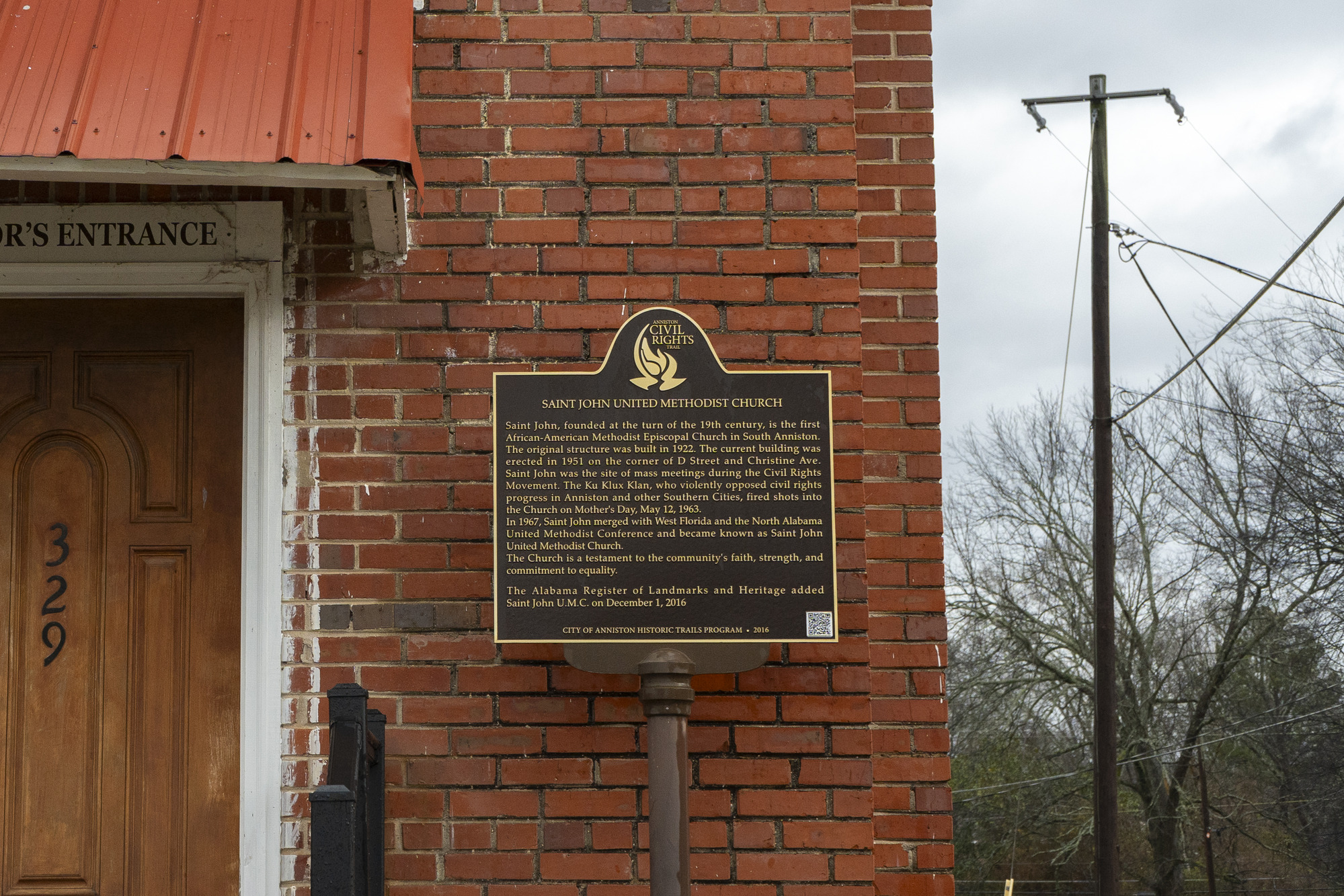 A metal marker saying Saint John United Methodist Church Anniston Civil Rights Trail. The sign is located to the right of a wooden door of the brick church. 