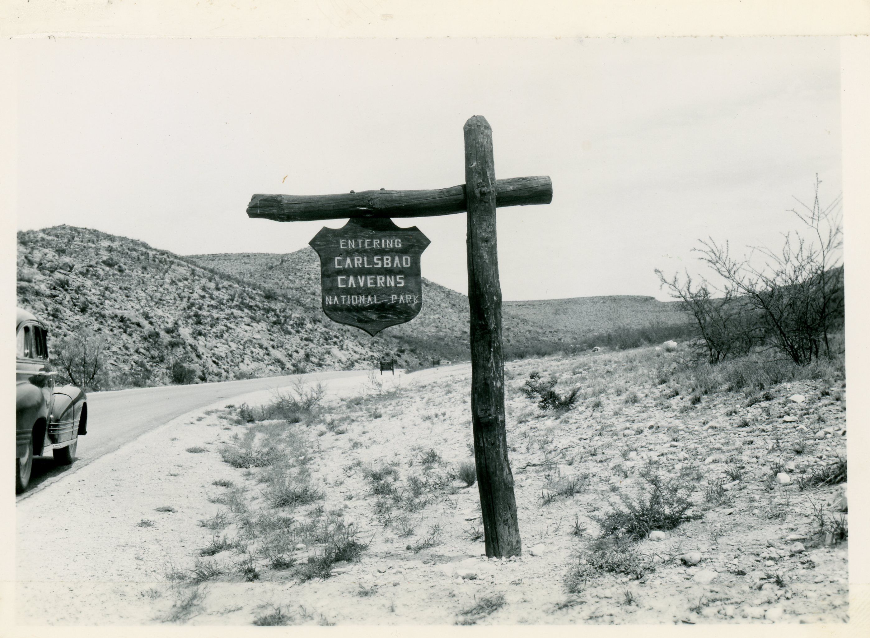 A black and white photograph of a wooden sign constructed on the side of the road, welcoming visitors to Carlsbad Caverns National Park.