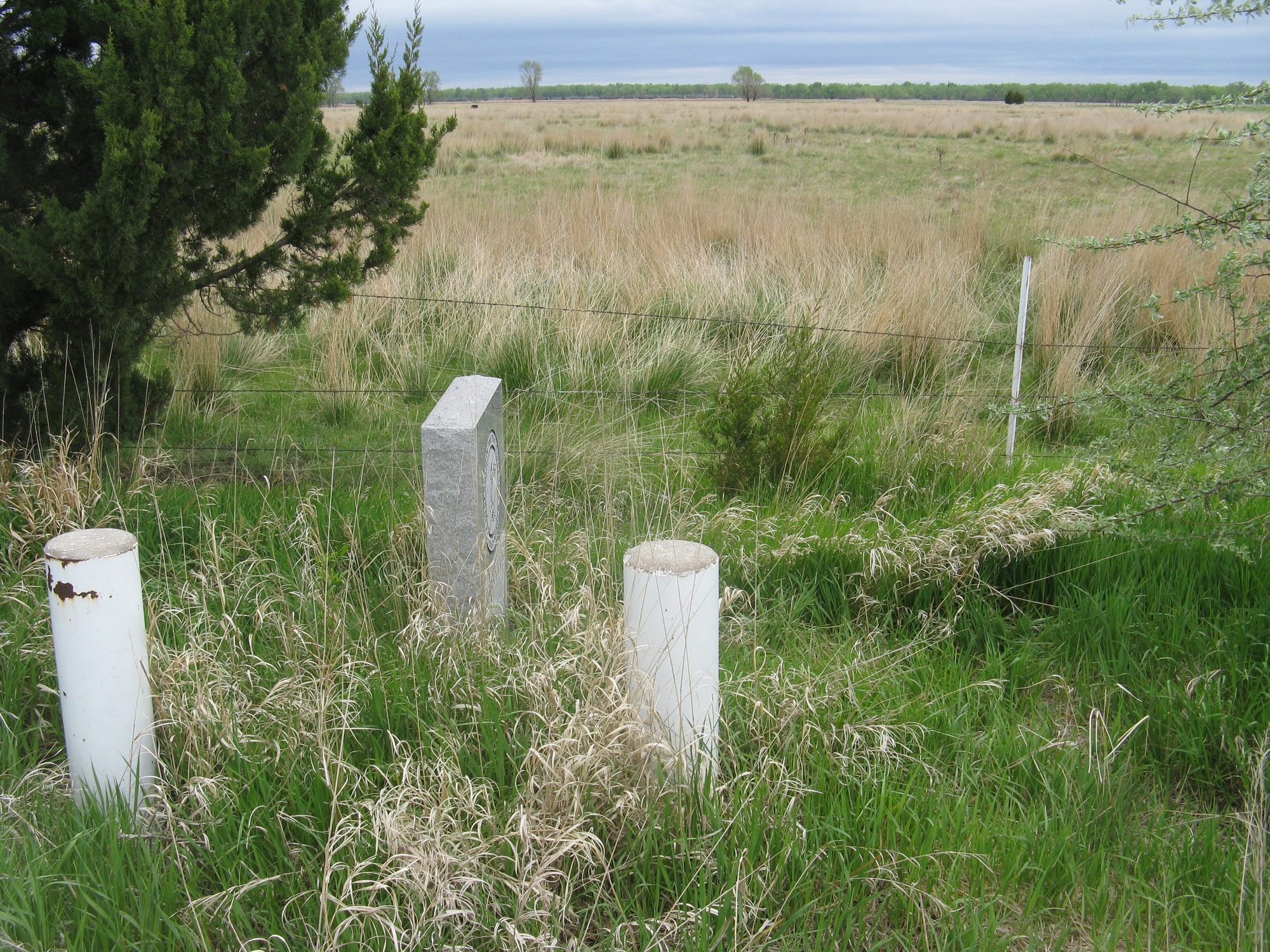 A stone marker and white concrete poles next to an abandoned farm field mark the location of Gilman's Pony Express Station