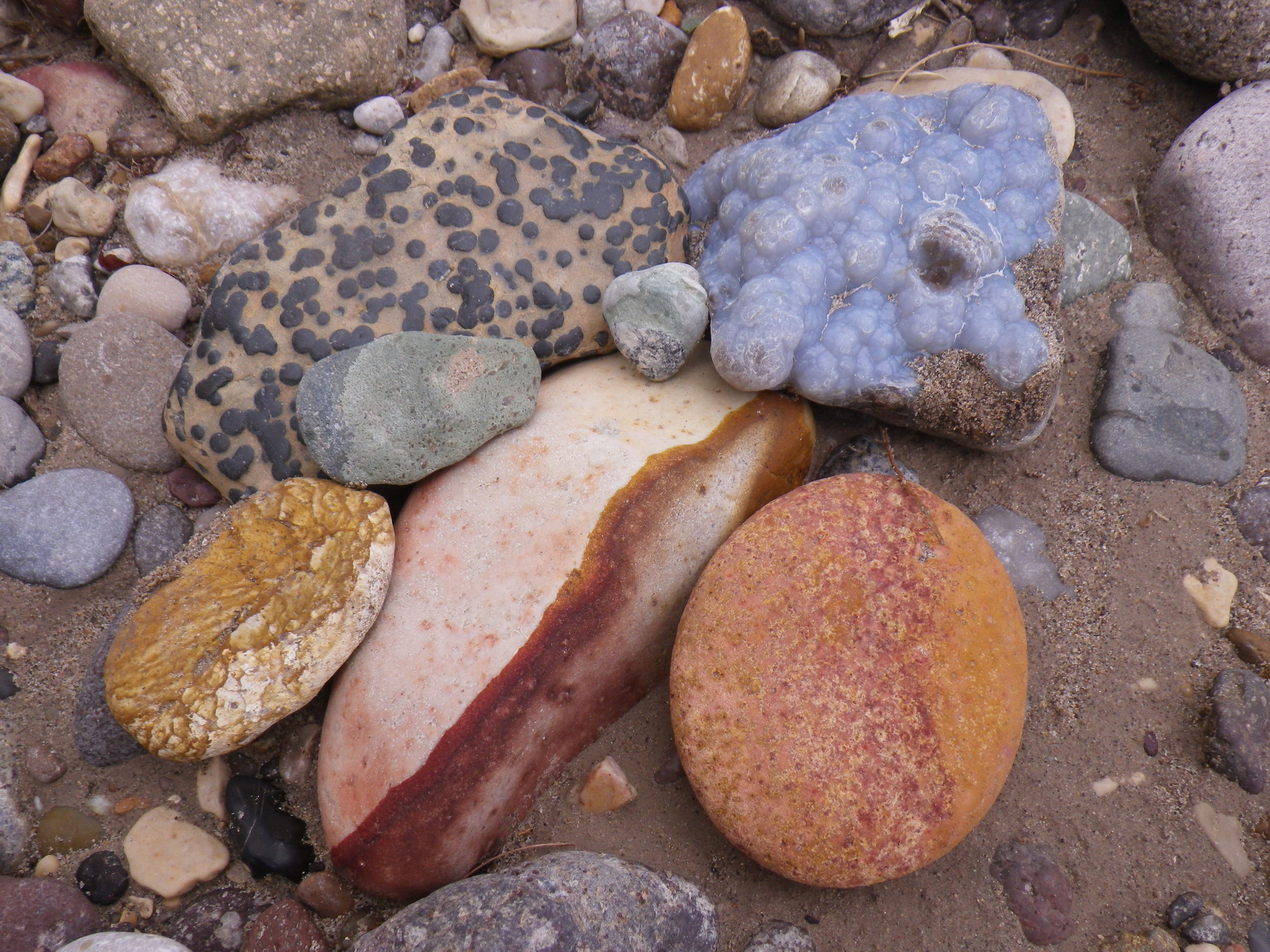 A collection of river rocks stacked on a bar of wet sand. Rocks include: a beige rock with black speckles; lavender-colored calcite; a round rock that changes colors from orange to red; a long, pink rock with a dark red band running laterally; and a pale yellow rock with dark yellow veins.