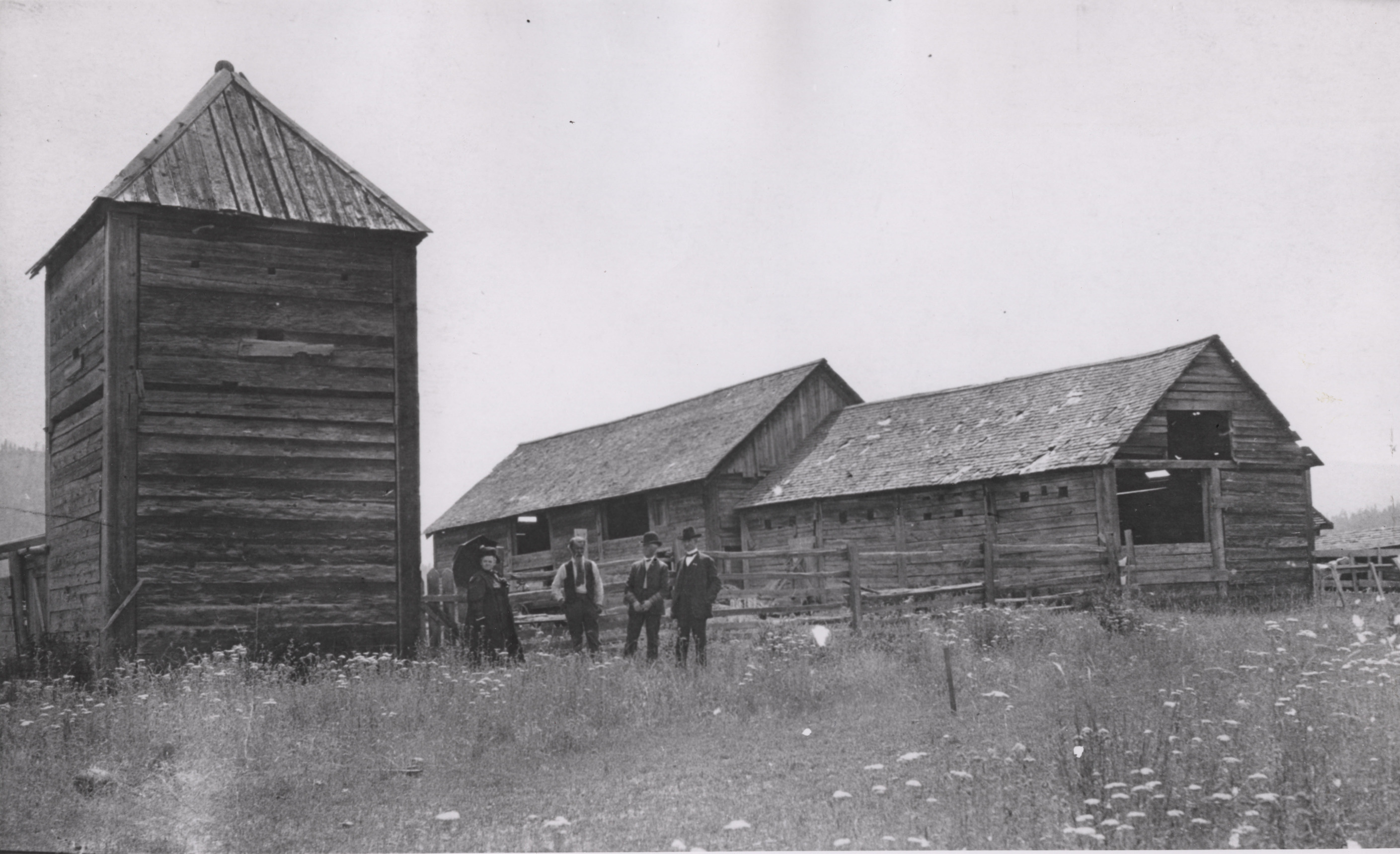 Black and white photograph of three men and a woman standing in front of several dilapidated wooden buildings