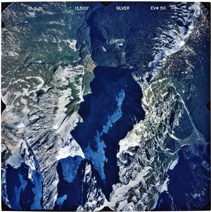 Color aerial photograph of glacier, snowfields, and alpine lake on mountain peak with forested valleys below.