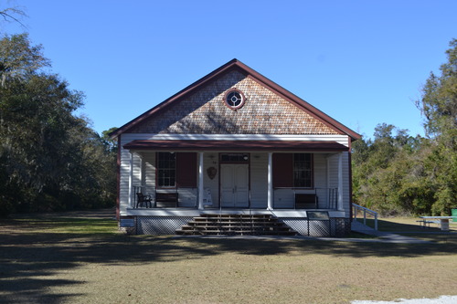 The white single-story building with a long front porch, red trim and shutters. The building has a main staircase of five steps with railings along the center steps at the front of the building. The front double door is at the center of the porch. Windows are on either side of the door and there are benches underneath each window. 