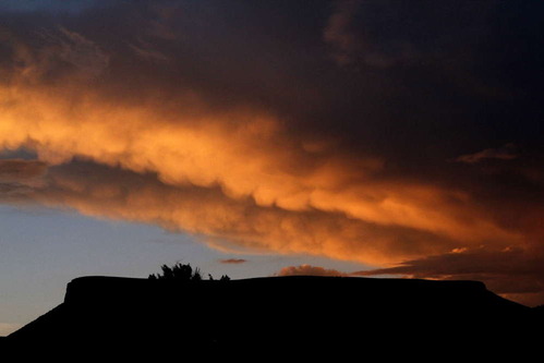 Clouds illuminated with orange light at sunset are cast over a silhouetted mesa.