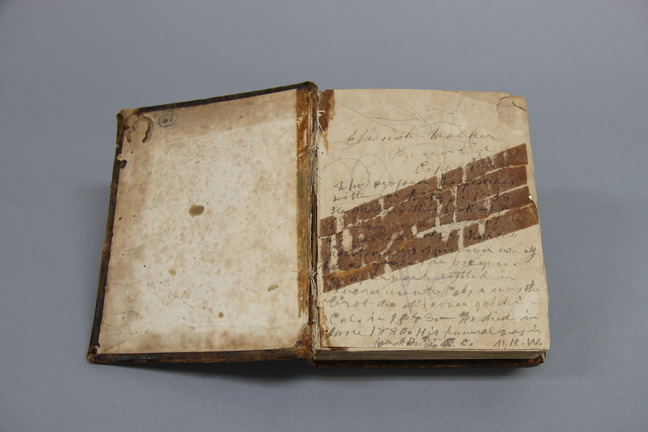 An old book with the front cover open, pages yellowed and speckled with age and spine cracking. The first page is filled with a handwritten note. The name Elkanah Walker can be made out.