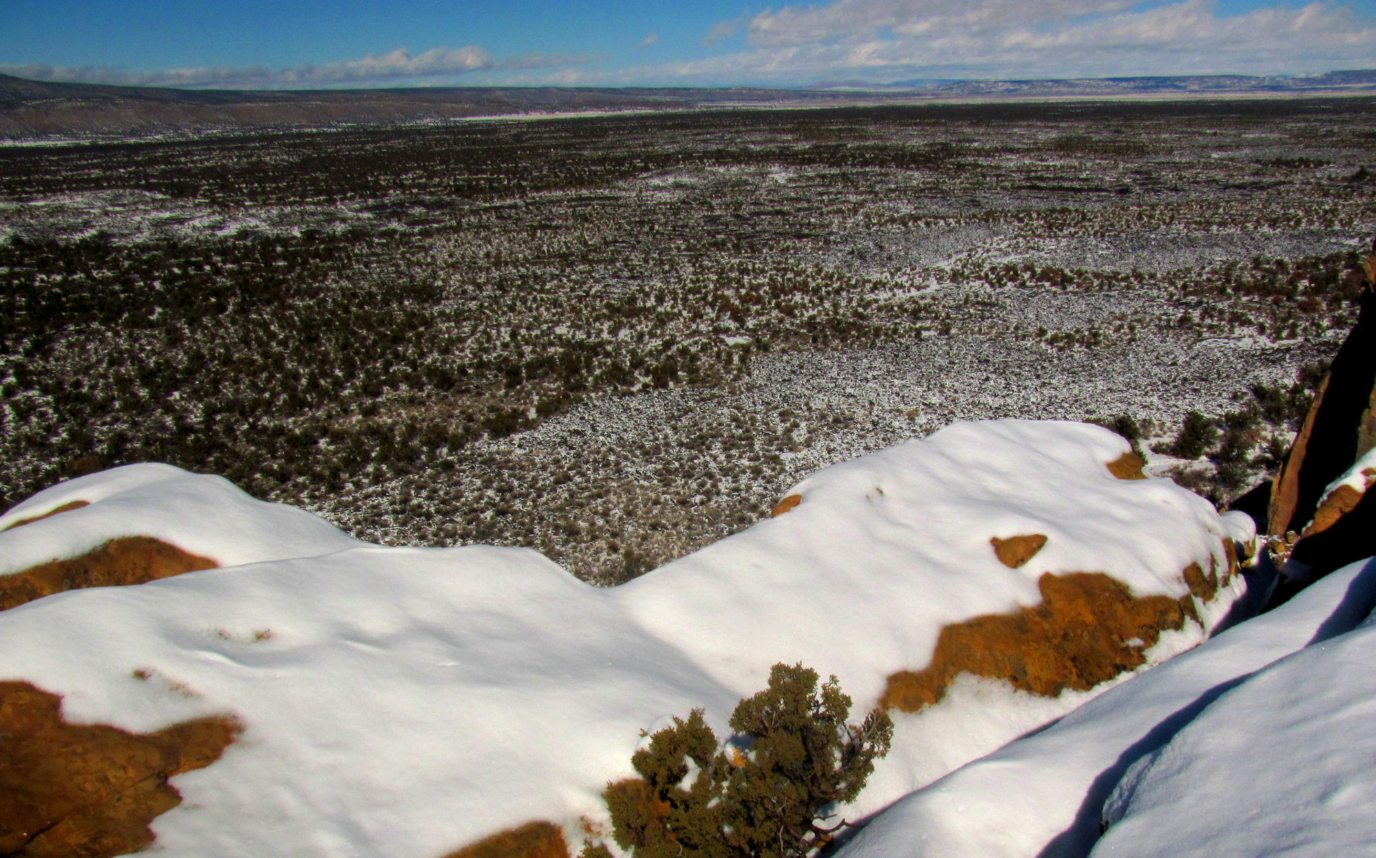 The edge of a yellow sandstone cliff peeks out from under a thin layer of snow.  A vast, rugged landscape of black rock also dusted with snow fills the landscape below and beyond it.  Clouds dot the blue sky above.