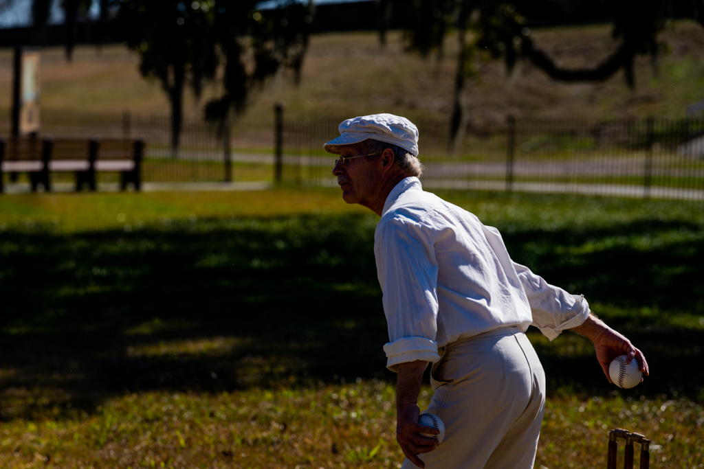 A man wearing a white shirt and white pants extends his arm behind him and holds a ball.