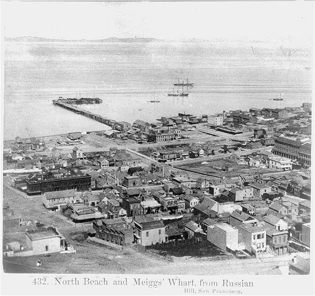 Black and white image of a birds eye view of San Francisco, California, looking towards North Beach from Russian Hill. Buildings are in the forefront, with the water and a few ships in the back.