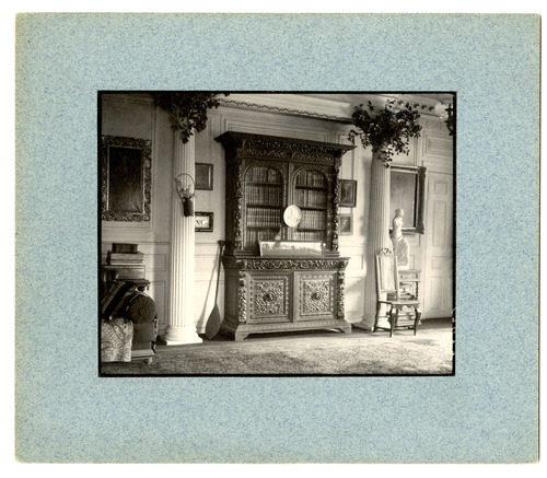 Black and white photograph of Renaissance revival book cases between Greek columns. Mounted on blue paper.