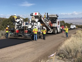Heavy equipment and workers laying new asphalt on road on left side and dirt underlayment on right side of road.