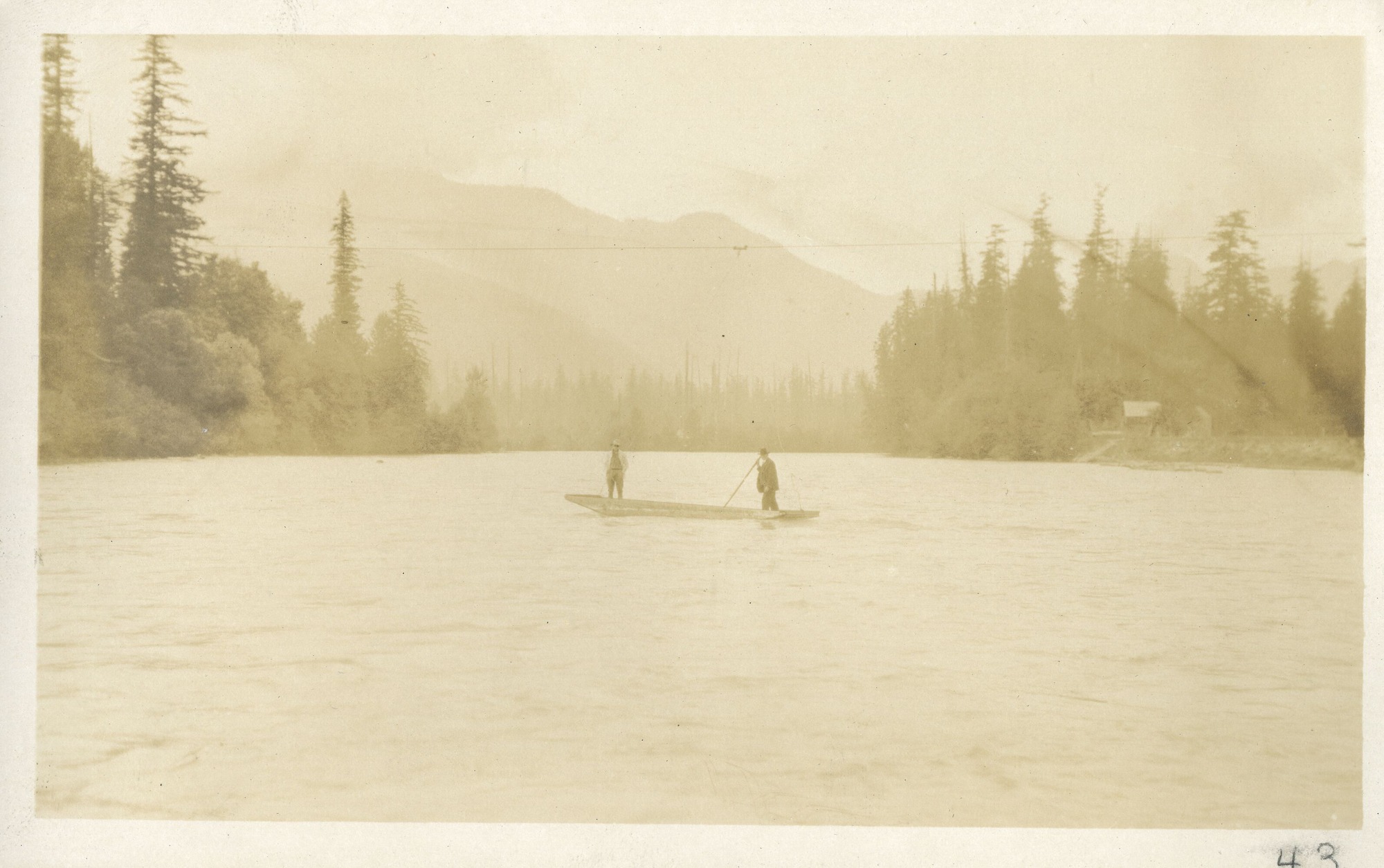 Two men standing in a boat on a river, cabin on the right bank in the distance.