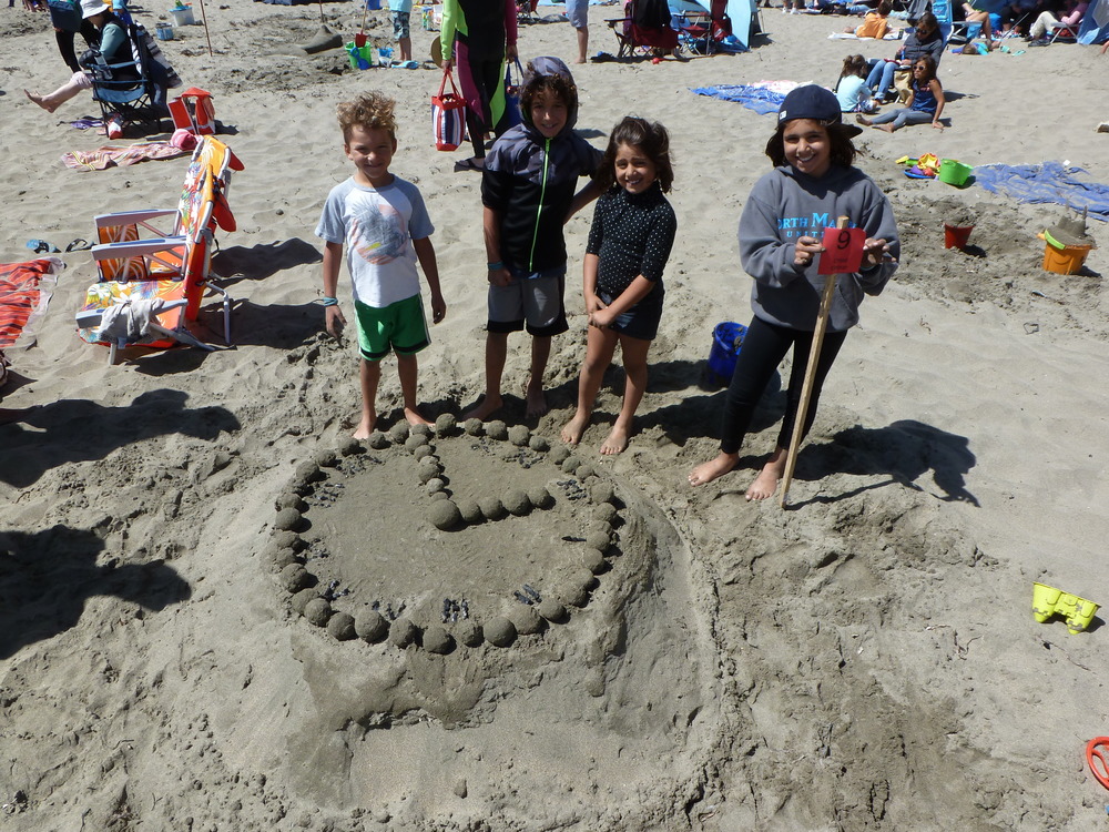 Four children standing behind a sand sculpture of a clock.