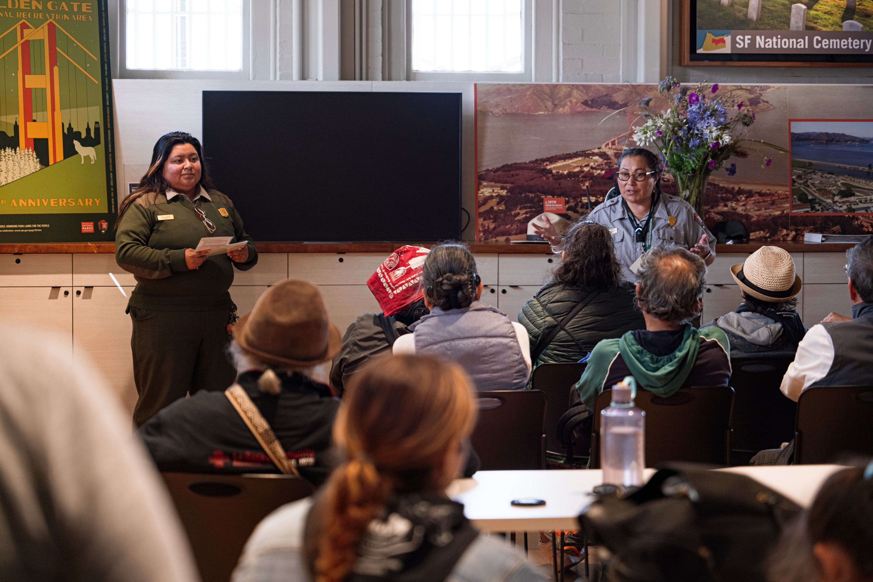 Two NPS employees in uniform present in front of a group of seated people inside of a building
