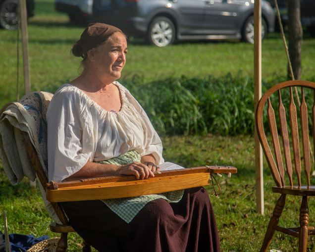 Woman seated in chair playing a dulcimer in period frontier clothing.