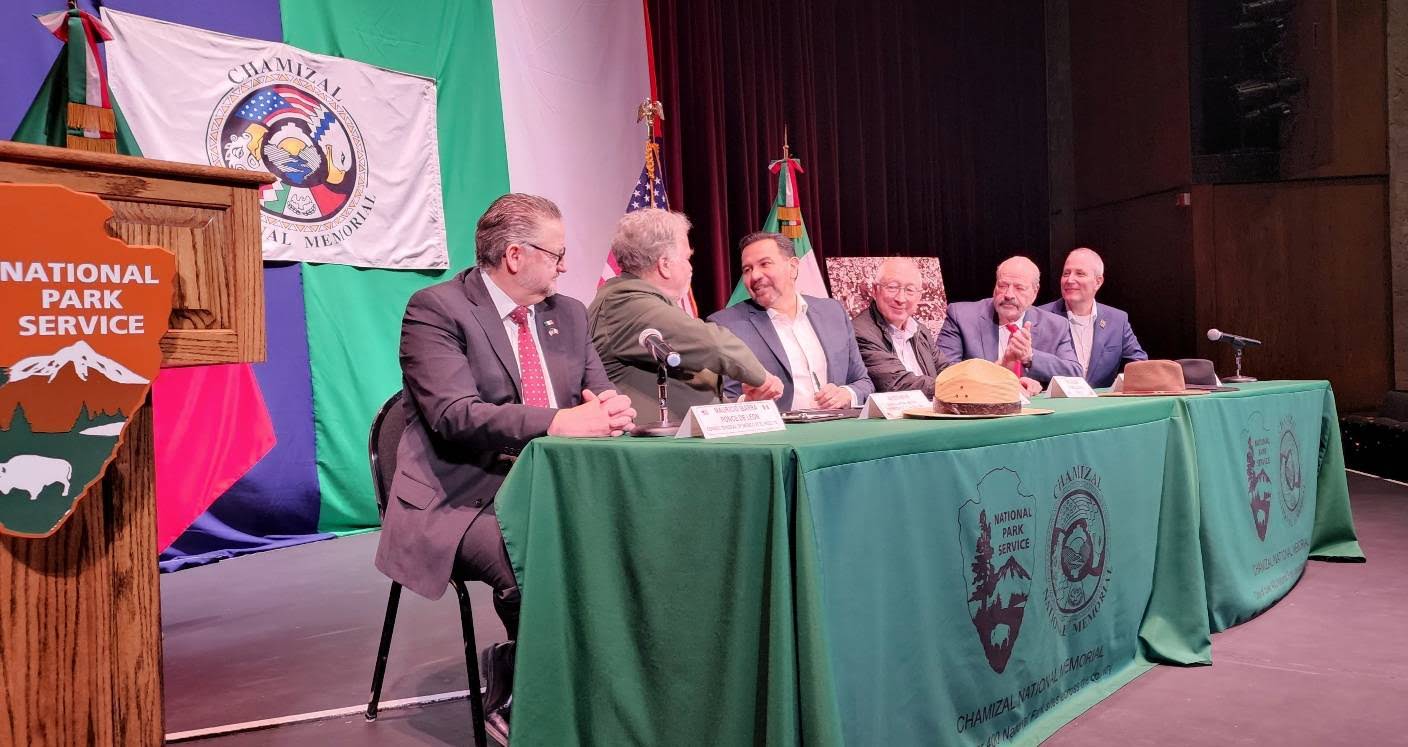 Six men seated at a table covered with the emblems of two protected area agencies, with flags of each entity in the background.