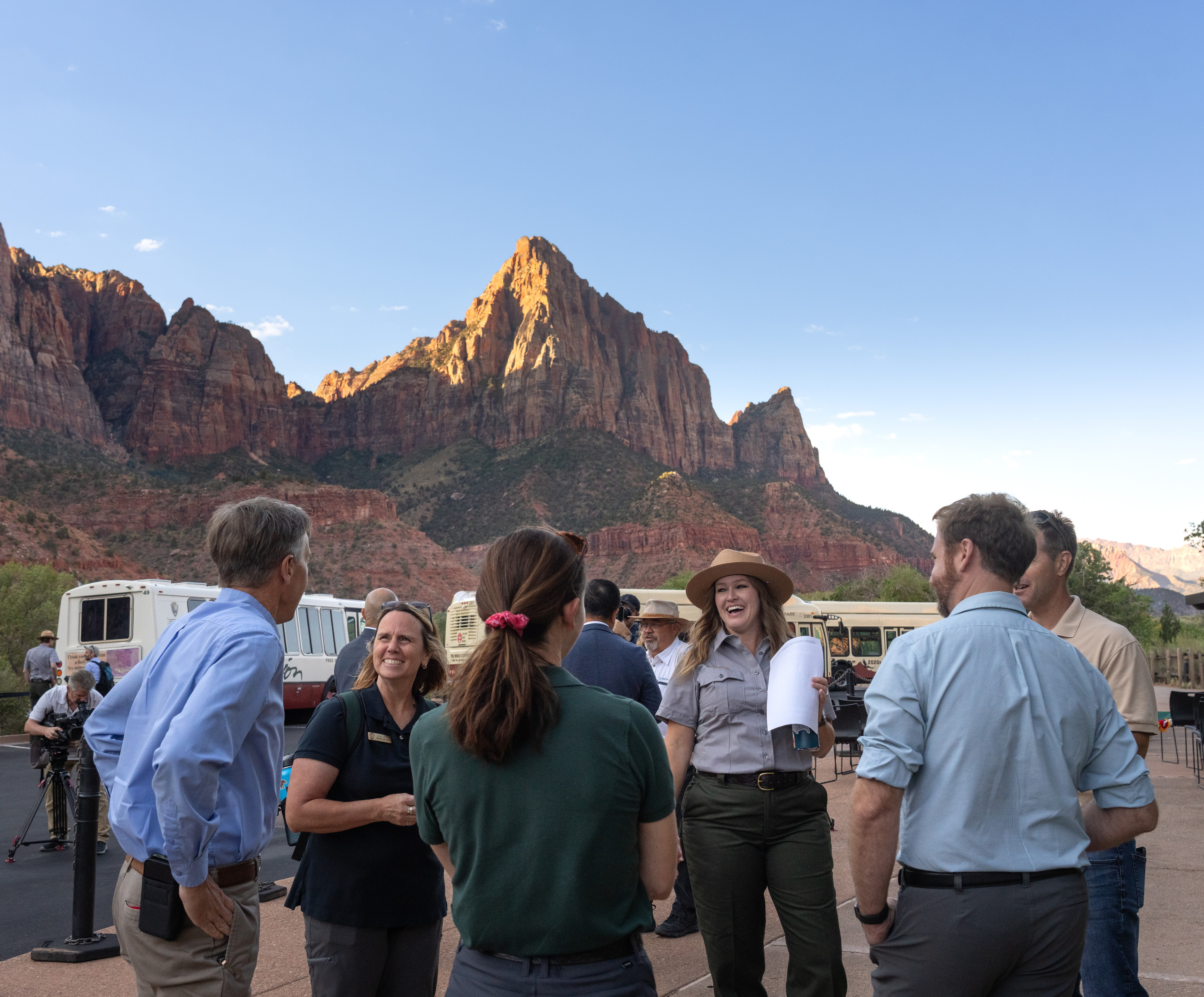 A crowd of people stand in front of the red, sandstone Watchman formation with several shuttle buses visible behind them. 