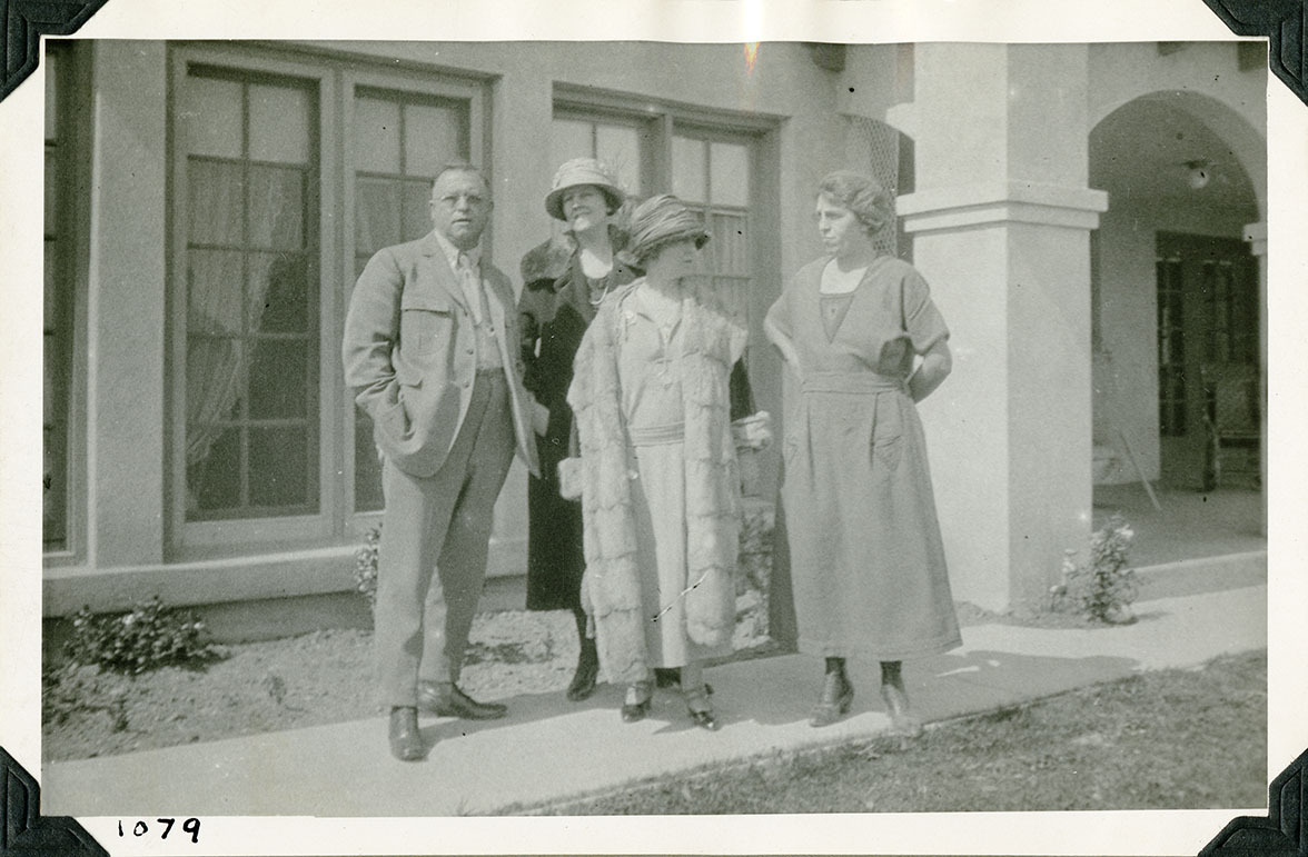 This is an historic black and white photograph from the Scotty's Castle Historic Photograph Collection, Death Valley National Park of four people in front of a large Spanish style house. Man in suit with woman in heavy coat. Another woman with 1920s garb and heavy coat. Woman on right in plan dress. Number in black ink in lower left corner.