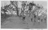 The image is in black and white. A group of six men and children span from the center of the image to the right edge. One man, the man towards the center of the photo, is putting. His back is facing the camera. The rest of the group watches him. Three of the men watching the golfer are identified. From left to right after the boy standing towards the center of the photo: Dick, Jack, and Dad (Channing) Lovejoy. The dirt surface the group is on is smooth. In the background, a cluster of koa trees spreads from the left end to the right end of the photo. A label at the bottom of the image in the white border reads, 