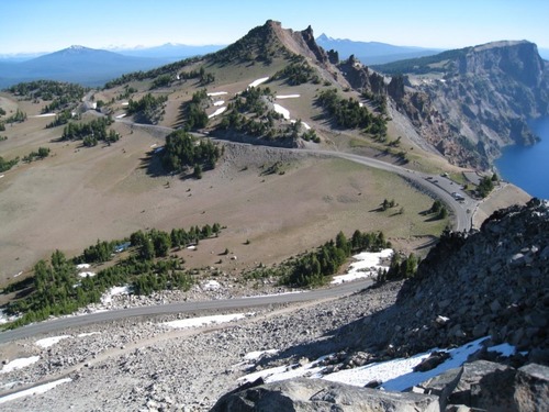 A high-angle view shows a road winding into the distance around peaks, dropping to the lake on the right.