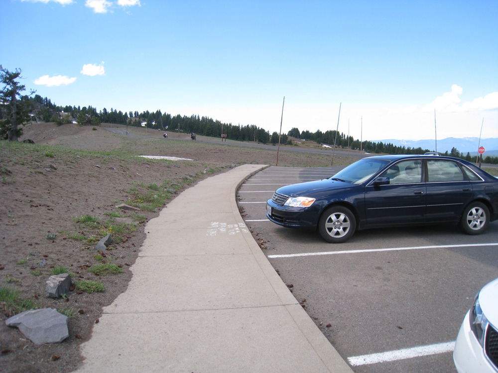 Several cars are parked near a curb in an overlook parking log, surrounded by open landscape and wide views.
