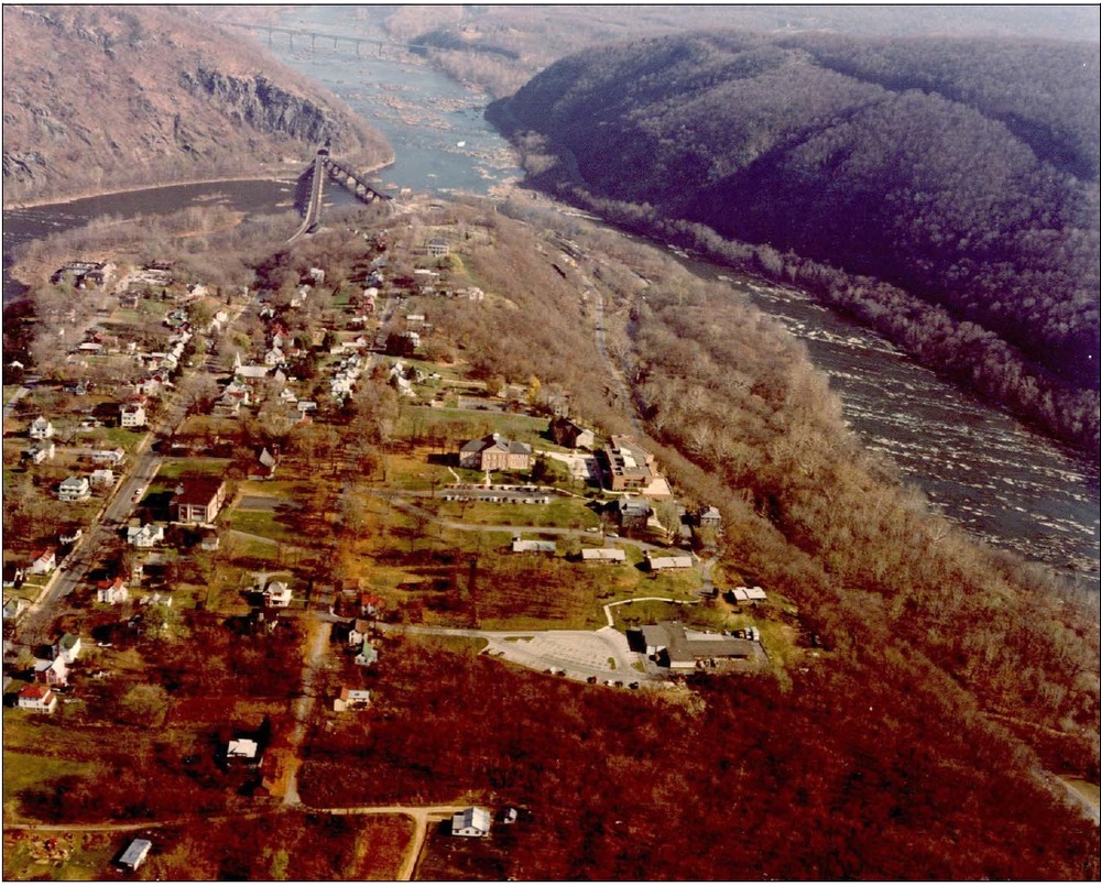 Aerial view shows sloping land between two rivers, dotted with buildings, trees, and roads.
