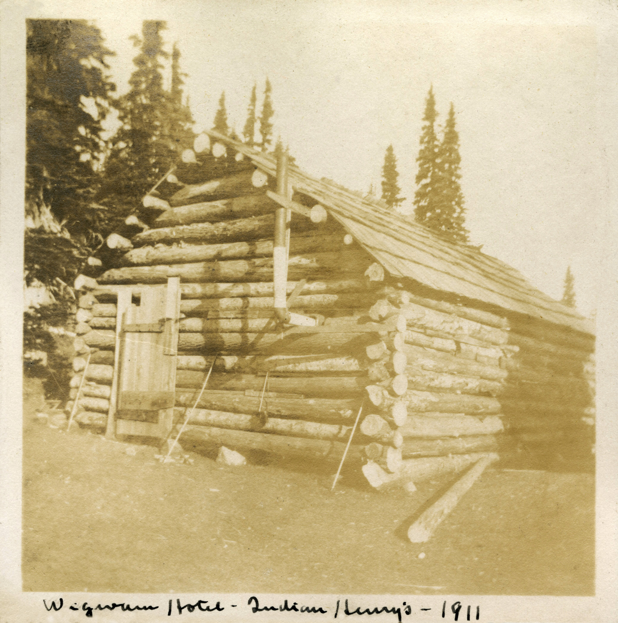 Historic photo of a roughly constructed log cabin with one door and no windows with a metal fireplace vent sticking out of the side. 
