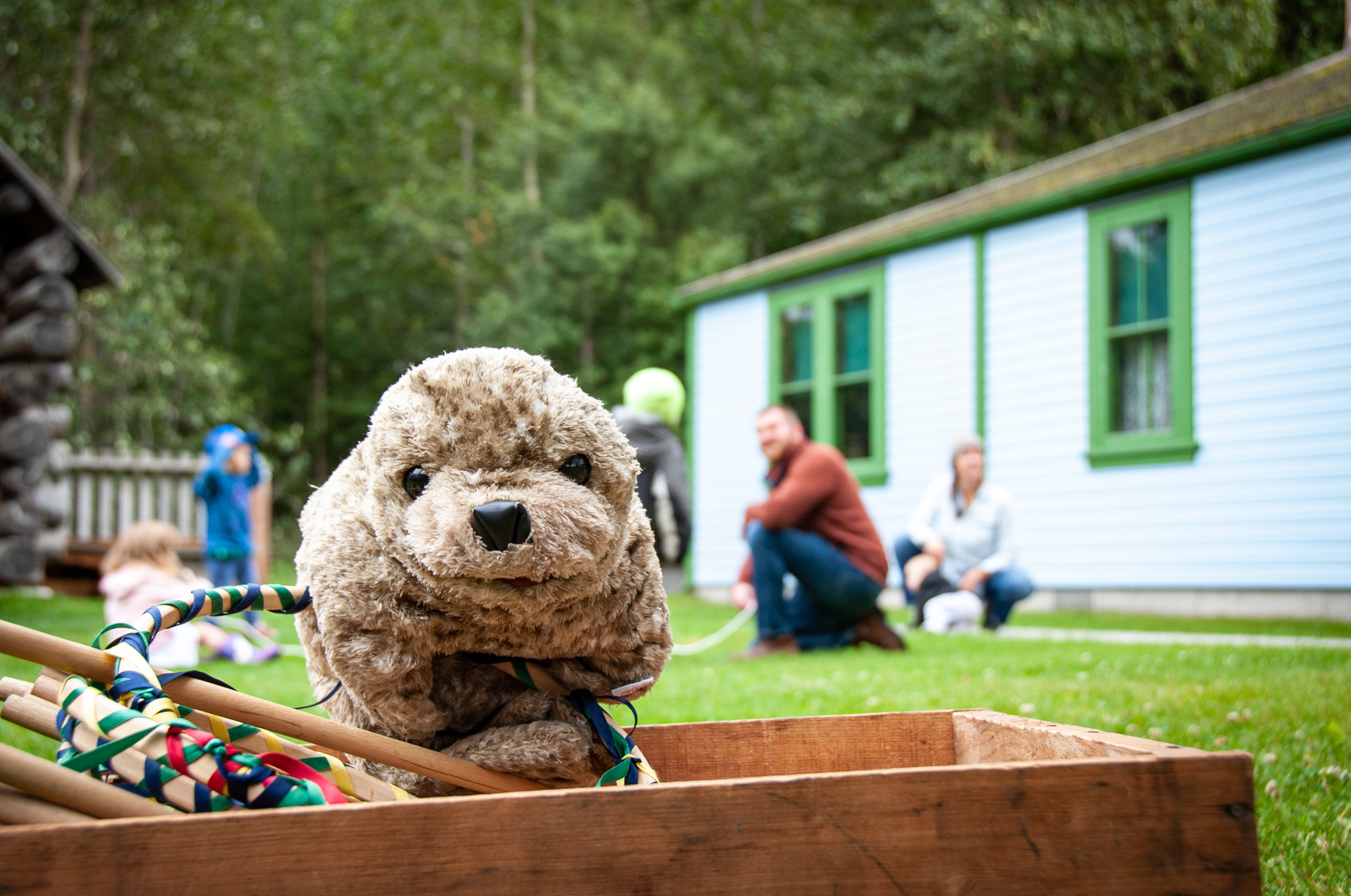 A stuffed toy otter in a box, with blurred children playing in the background