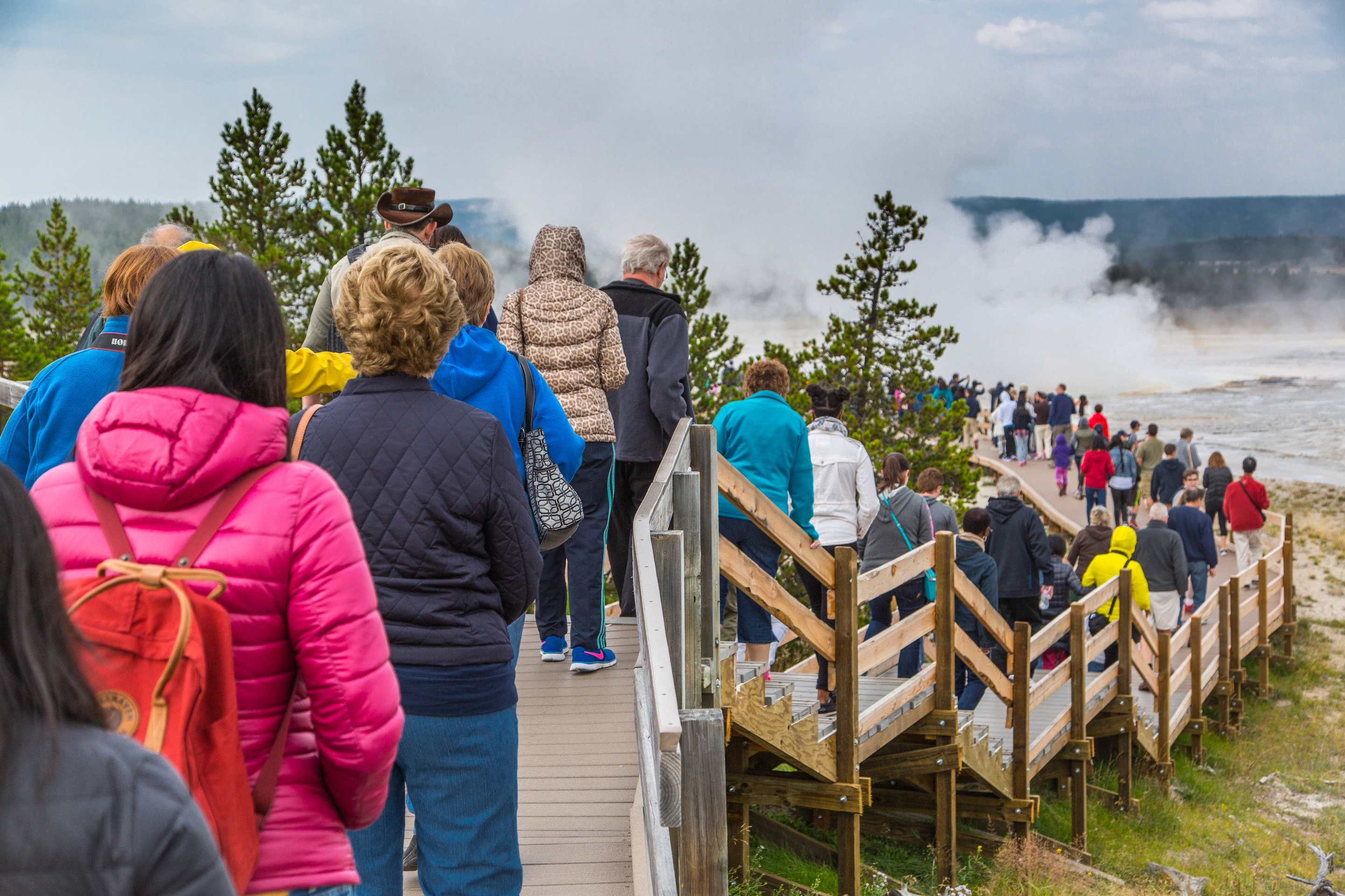 A boardwalk which approaches a steamy geyser is full of people.